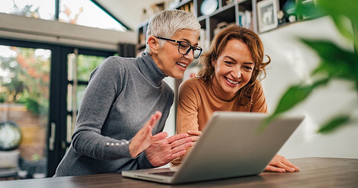 Two women are smiling and engaging with a laptop, sharing a joyful moment in a cozy indoor setting