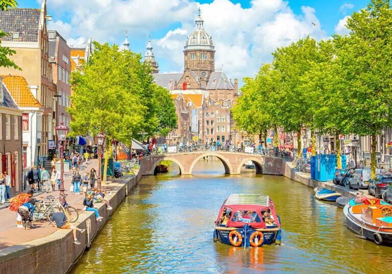 Scenic Amsterdam canal with a tour boat, historic buildings and the St. Nicholas Church in the background, representing the financial and tax planning considerations for American expats moving to the Netherlands