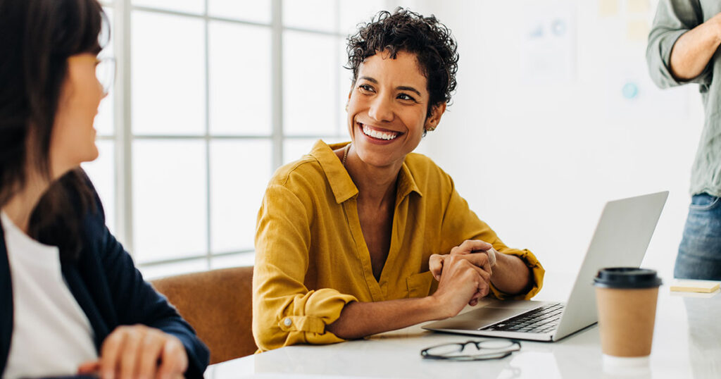 Two colleagues smiling and discussing retirement planning over a laptop at an office desk.