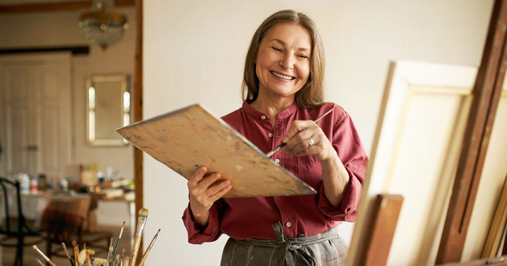 Happy retired woman smiling while painting on a canvas in her home art studio, representing how today's retirees are structuring a purposeful and fulfilling retirement lifestyle