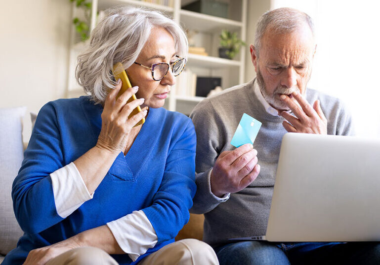 Concerned older couple at home on the phone and holding a credit card while reviewing a laptop, representing the risk seniors face from common financial scams and identity theft