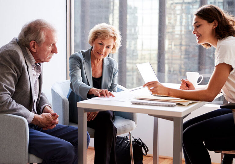 Older couple reviewing documents with a young female financial advisor at a table in a modern high-rise office, representing a conversation about investment diversification and portfolio planning