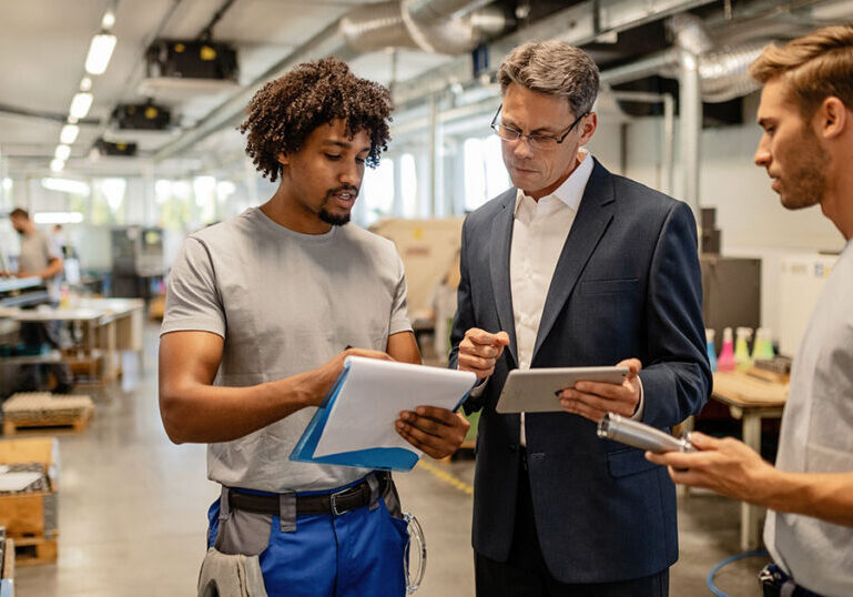 Business manager reviewing documents on a tablet with two workers on a manufacturing floor, representing the hidden operational costs of scaling a small business from startup to mid-market