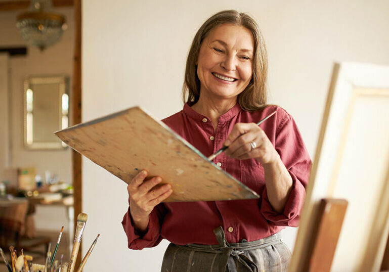 Happy retired woman smiling while painting on a canvas in her home art studio, representing how today's retirees are structuring a purposeful and fulfilling retirement lifestyle