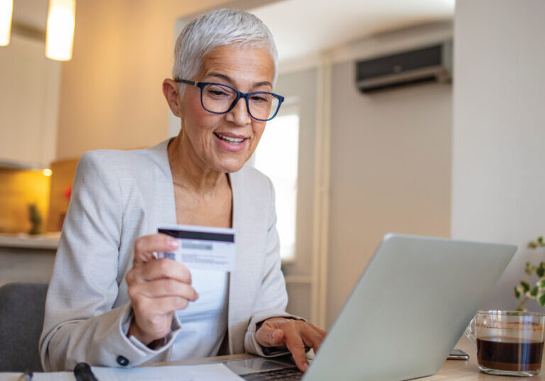 Woman reviewing financial information on a laptop and holding a credit card at her kitchen table