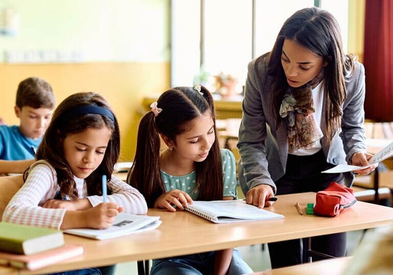 Elementary school teacher supporting two young students in class, symbolizing how outsourced school finance services help K‑12 and charter schools focus resources on teaching and learning