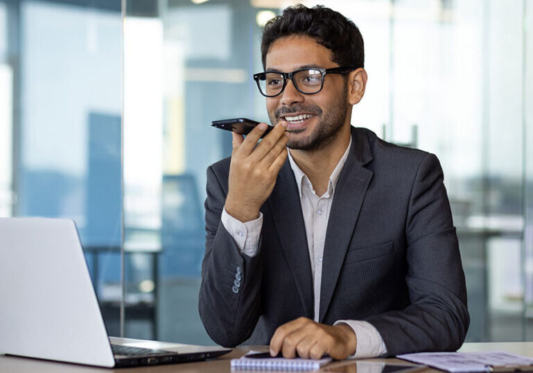 Small business owner speaking into a smartphone at his desk beside a laptop, reflecting the risks of relying on AI for accounting decisions without professional validation