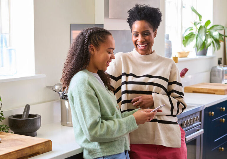 Parent and teenage child standing together in a bright kitchen, talking while looking at a smartphone.