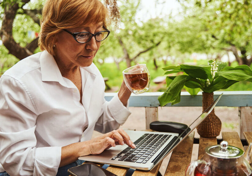 Pretty mature lady is sitting on terrace in countryside and typing on notebook while drinking tea