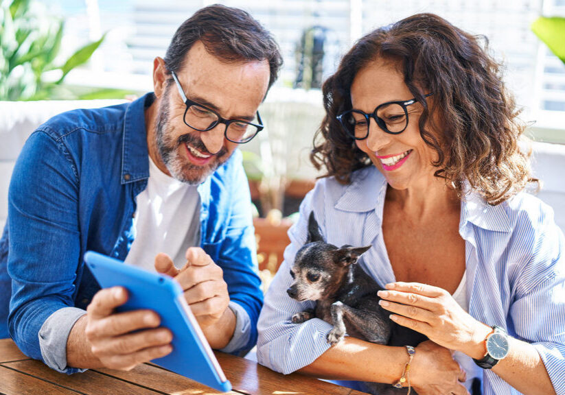 Middle age hispanic couple using touchpad sitting on table with dog at terrace