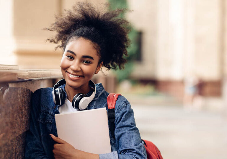 Happy African American Student Girl Holding Books Posing Standing Near University Building Outside, Smiling To Camera. Modern Education, College Study Concept